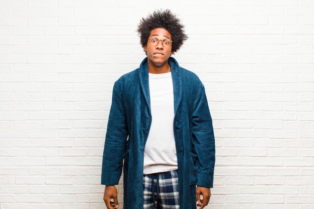 Young black man wearing pajamas with gown feeling clueless, confused and uncertain about which option to pick, trying to solve the problem against brick wall