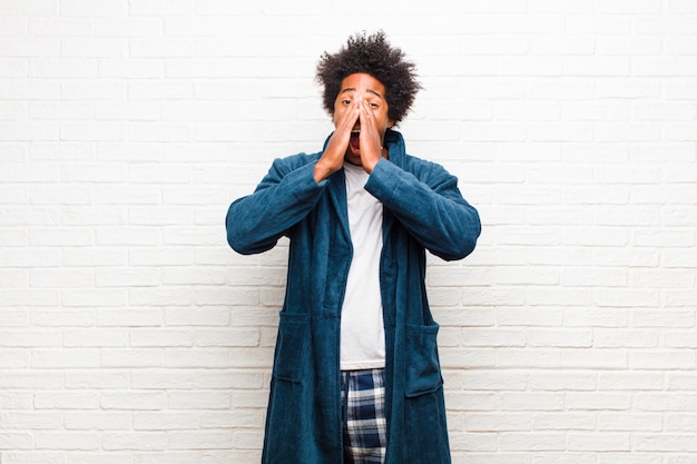 Young black man wearing pajamas with gown feeling happy, excited and positive, giving a big shout out with hands next to mouth, calling out against brick wall