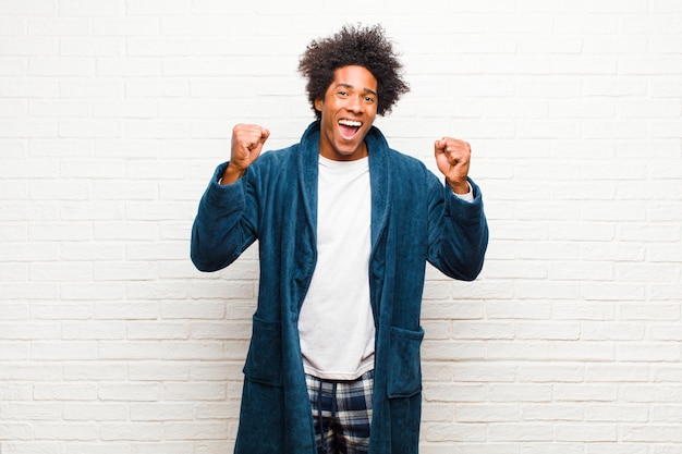 Young black man wearing pajamas with gown feeling happy, surprised and proud, shouting and celebrating success with a big smile on brick wall