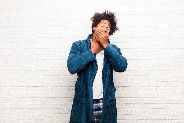 Young black man wearing pajamas with gown feeling ill with a sore throat and flu symptoms, coughing with mouth covered against brick wall