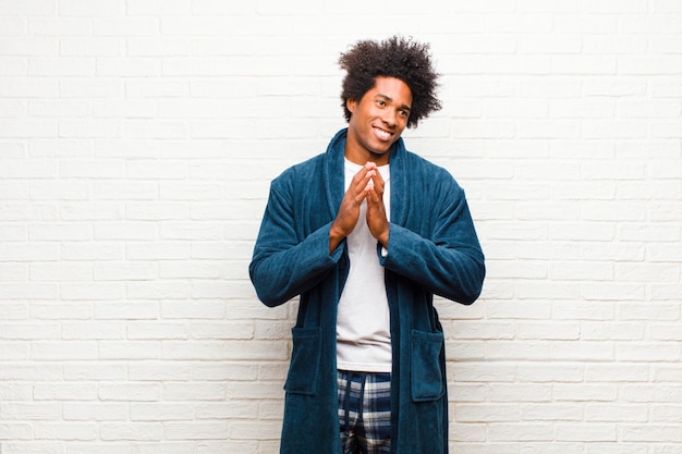 Young black man wearing pajamas with gown feeling proud mischievous and arrogant while scheming an evil plan or thinking of a trick against brick wall