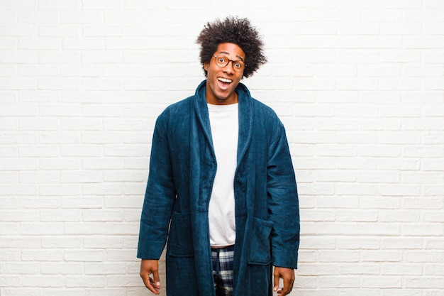 Photo young black man wearing pajamas with gown looking happy and pleasantly surprised excited with a fascinated and shocked expression against brick wall