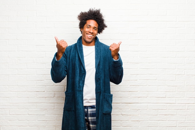 Young black man wearing pajamas with gown smiling joyfully and looking happy, feeling carefree and positive with both thumbs up against brick wall