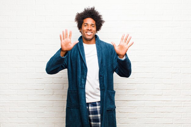 Young black man wearing pajamas with gown smiling and looking friendly, showing number tententh with hand forward, counting down  brick wall