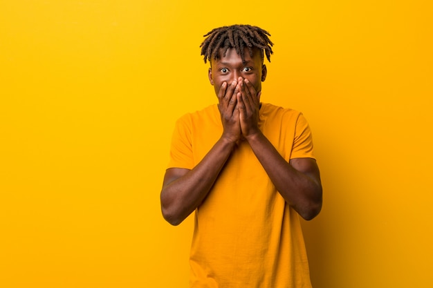 Young black man wearing rastas over yellow background laughing about something, covering mouth with hands.