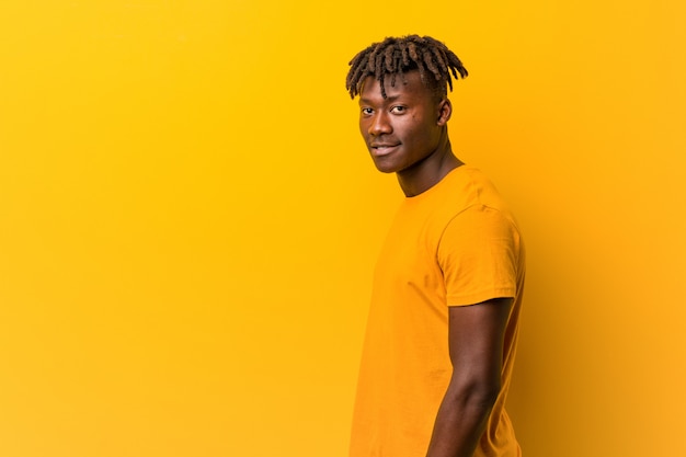 Young black man wearing rastas over yellow wall looks aside smiling, cheerful and pleasant