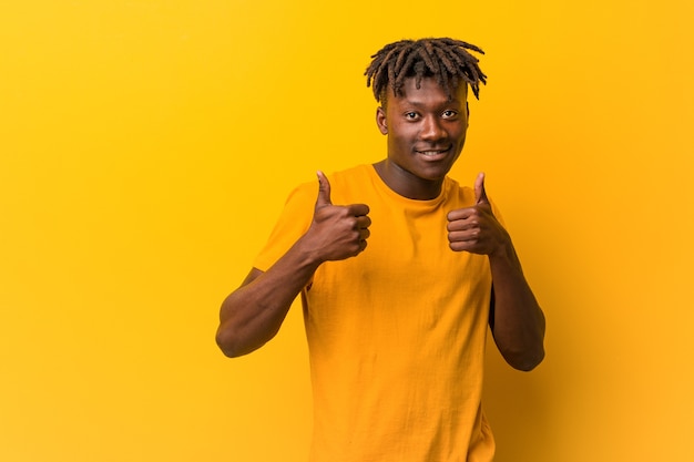 Young black man wearing rastas over yellow wall raising both thumbs up, smiling and confident.