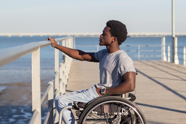 Молодой темнокожий мужчина в инвалидной коляске смотрит на морской пейзаж. Вид сбоку на уверенного в себе стильного парня с параличом нижних конечностей, кладущего руку на перила и наслаждающегося видом на море. Средний выстрел. Инвалидность, концепция отношения.