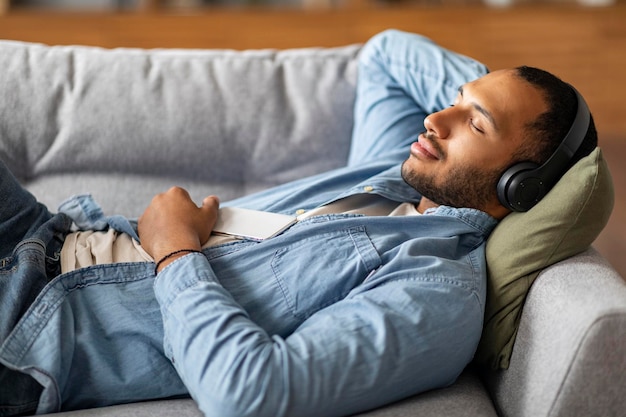 Young Black Man In Wireless Headphones Relaxing On Couch With Smartphone