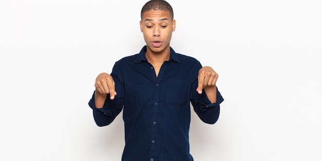 Photo young black man with open mouth pointing downwards with both hands, looking shocked, amazed and surprised