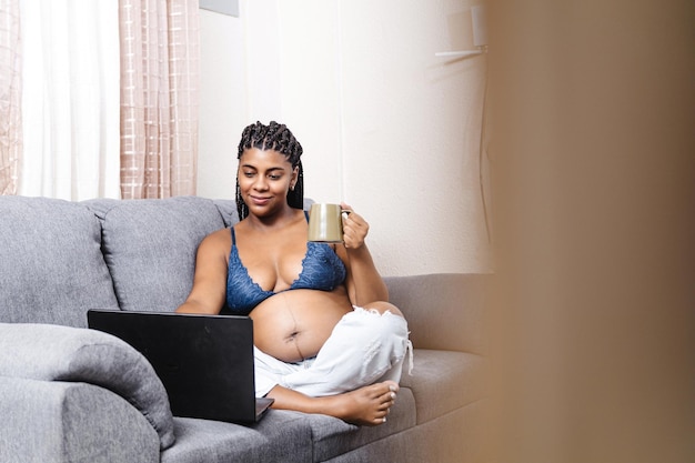 Young black pregnant woman sitting holding a cup using a laptop