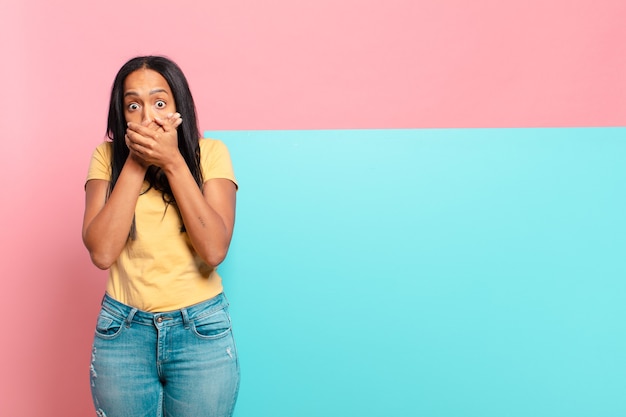 Giovane donna di colore che copre la bocca con le mani con un'espressione scioccata e sorpresa, mantenendo un segreto o dicendo oops. copia il concetto di spazio