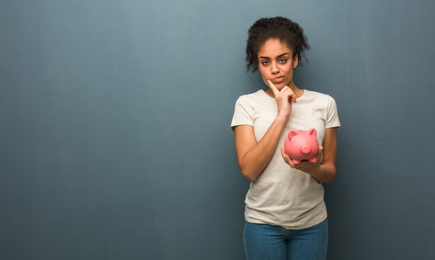 Young black woman doubting and confused. She is holding a piggy bank.