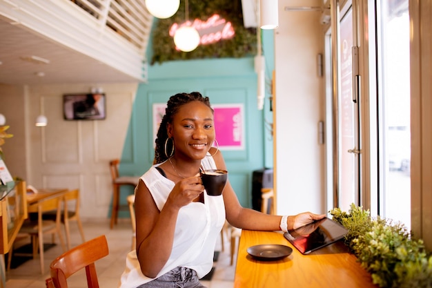 Giovane donna di colore che beve caffè mentre guarda la tavoletta digitale nella caffetteria