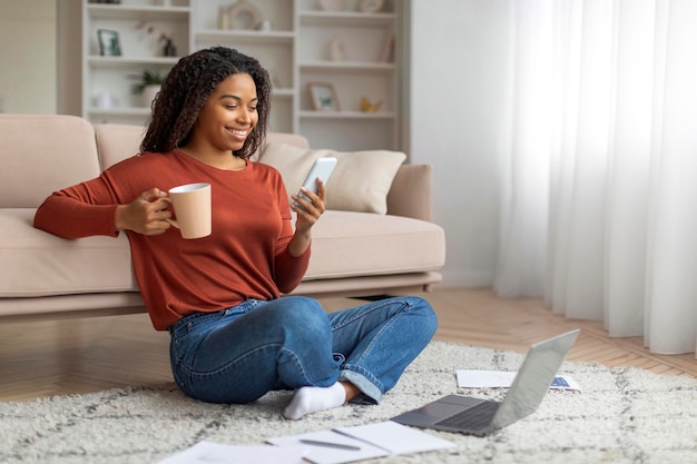 Young black woman enjoying coffee and using smartphone indoors