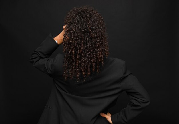 Young black woman feeling clueless and confused, thinking a solution, with hand on hip and other on head, rear view against black wall