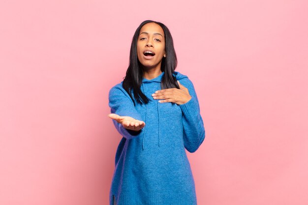 Young black woman feeling happy and in love, smiling with one hand next to heart and the other stretched up front