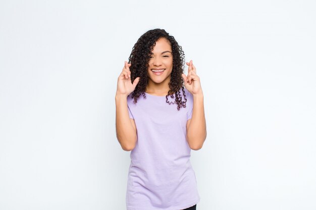 Young black woman feeling nervous and hopeful, crossing fingers, praying and hoping for good luck against white wall