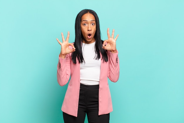 Young black woman feeling shocked, amazed and surprised, showing approval making okay sign with both hands. business concept