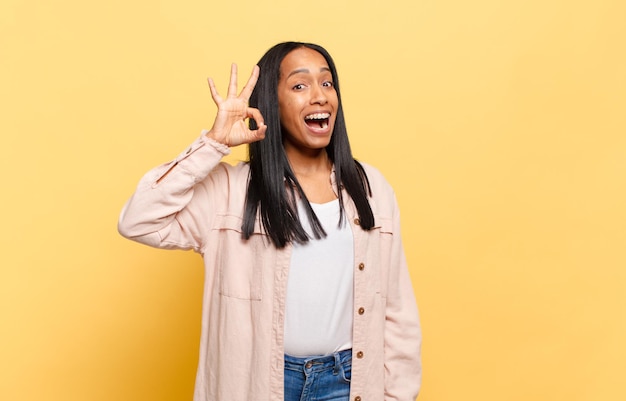 Young black woman feeling successful and satisfied, smiling with mouth wide open, making okay sign with hand