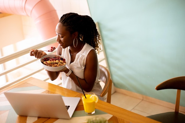 Giovane donna di colore che mangia una sana colazione mentre lavora al computer portatile nella caffetteria