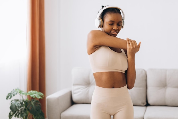 Young black woman in headphones listens to music and does workout at home in the living room. Home training.