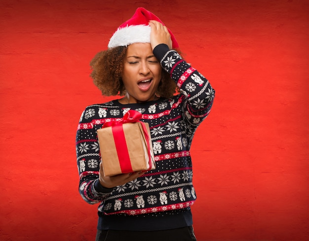 Young black woman holding a gift in christmas day forgetful, realize something