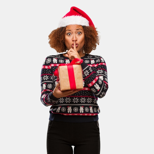 Young black woman holding a gift in christmas day keeping a secret or asking for silence