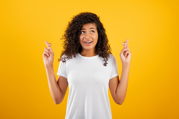 Young black woman hoping and praying with fingers crossed isolated over yellow