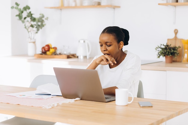 A young black woman is sitting in the kitchen working on a laptop, she is tired, yawns and wants to sleep.