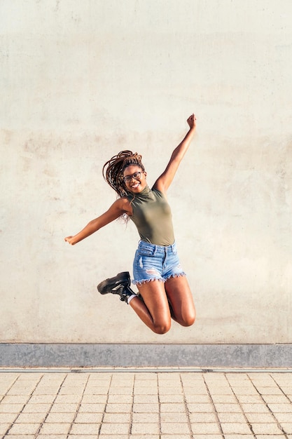 Foto la giovane donna nera salta felice e sorridente con un muro di cemento grigio sullo sfondo, concetto di gioventù e diversità razziale, copia spazio per il testo