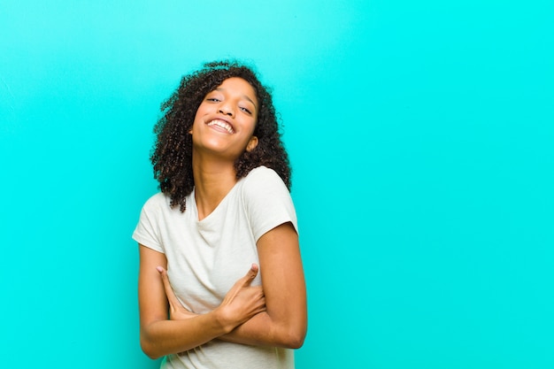 Young black woman laughing happily with arms crossed, with a relaxed, positive and satisfied pose on blue wall