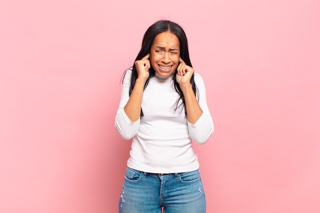 怒り、ストレス、イライラしているように見える若い黒人女性は、耳をつんざくような音、音、または大音量の音楽で両耳を覆っています