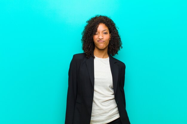 Young black woman looking happy and friendly, smiling and winking an eye at you with a positive attitude against blue wall