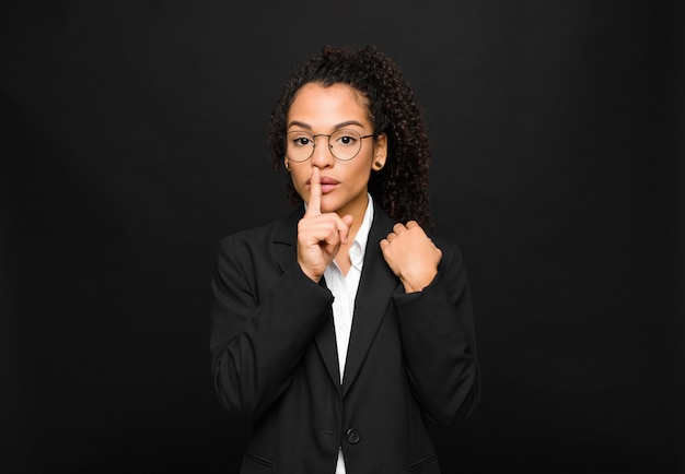 Young black woman looking serious and cross with finger pressed to lips demanding silence or quiet, keeping a secret against black wall