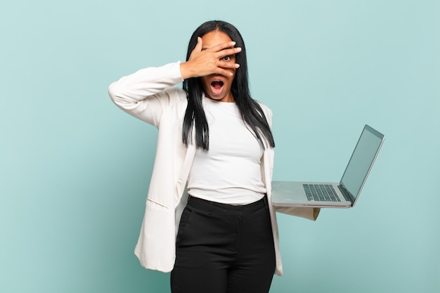 Young black woman looking shocked, scared or terrified, covering face with hand and peeking between fingers. laptop