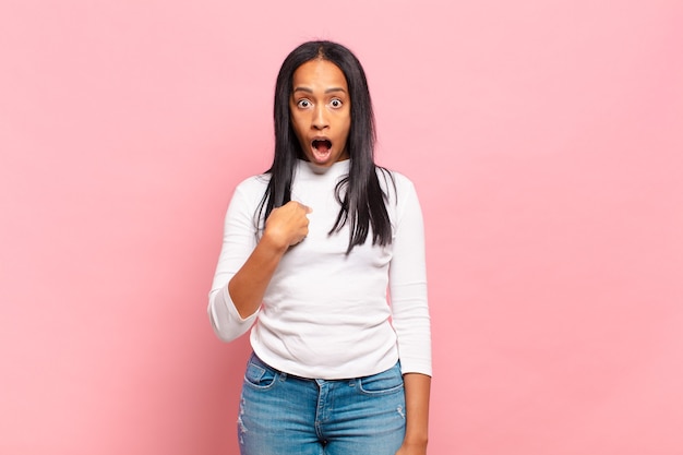 Young black woman looking shocked and surprised with mouth wide open, pointing to self
