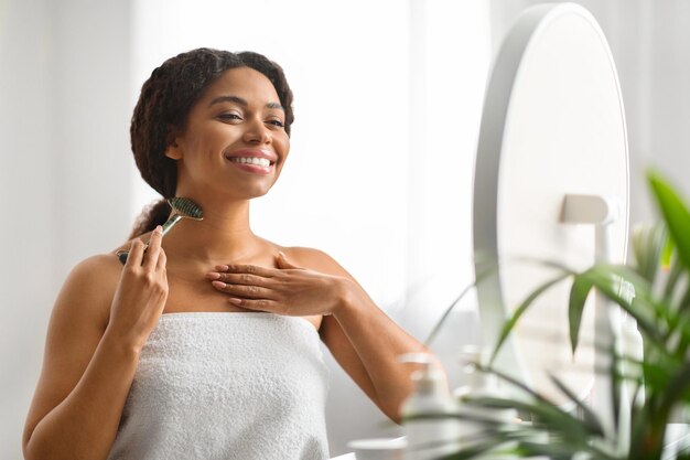 Photo young black woman massaging neck with greenstone jade roller near mirror