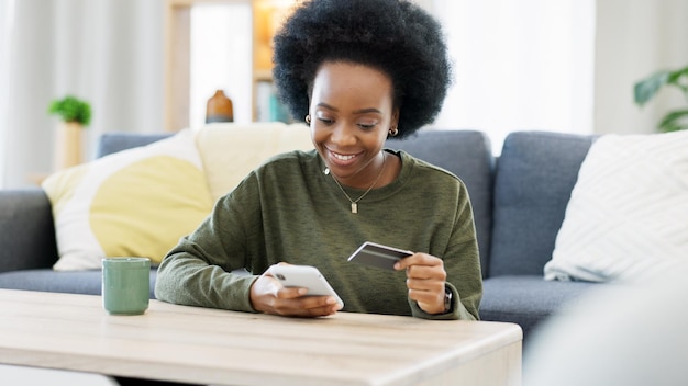 明るいリビング ルームで電話と銀行クレジット カードを使用してオンライン ショッピングをする若い黒人女性