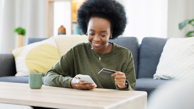 Young black woman shopping online using her phone and bank credit card in a bright living room Banking and managing assets and finance using convenient wireless technology from the comfort of home