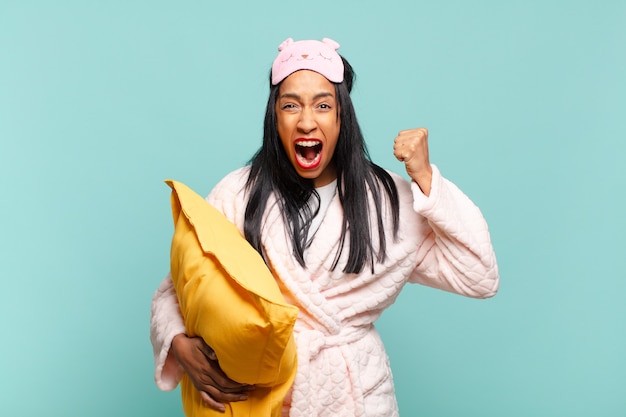 Young black woman shouting aggressively with an angry expression or with fists clenched celebrating success. pajamas concept