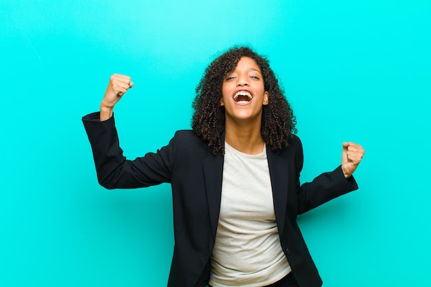Young black woman shouting triumphantly, looking like excited, happy and surprised winner, celebrating against blue wall