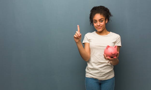 Young black woman showing number one. She is holding a piggy bank.