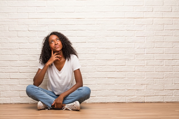 Young black woman sitting on a wooden floor doubting and confused, thinking of an idea or worried about something