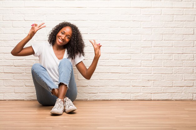 Young black woman sitting on wooden floor fun and happy, positive and natural, makes a gesture of victory, peace concept