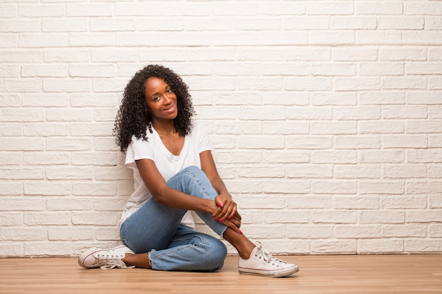 Young black woman sitting on a wooden floor with hands on hips, standing, relaxed and smiling