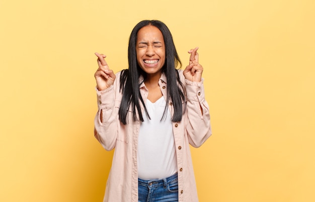 Photo young black woman smiling and anxiously crossing both fingers, feeling worried and wishing or hoping for good luck