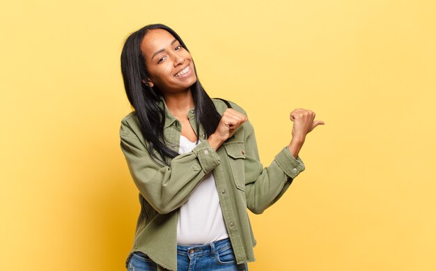 Young black woman smiling cheerfully and casually pointing to copy space on the side, feeling happy and satisfied