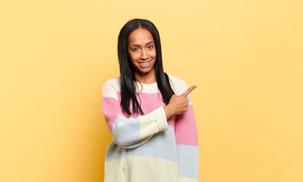 Young black woman smiling cheerfully, feeling happy and pointing to the side and upwards, showing object in copy space