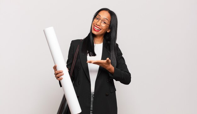 Young black woman smiling cheerfully, feeling happy and showing a concept in copy space with palm of hand. architect concept